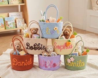 Könnte beinhalten: Sechs runde Körbe aus geflochtenem Seil in Orange, Beige, Hellblau, Gelb, Lila und Grün. Jeder Korb ist auf der Vorderseite mit einem personalisierten Namen in Schreibschrift bestickt und enthält kleine Plüschtiere wie Bären und Karotten.