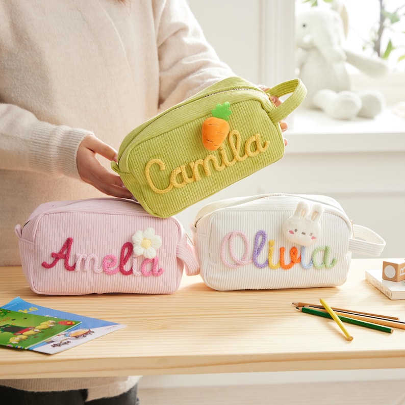 May include: Three rectangular corduroy pencil cases in light green, light pink, and white. The green case features the name Camila and a carrot patch. The pink case displays the name Amelia and a flower. The white case shows the name Olivia and a rabbit.