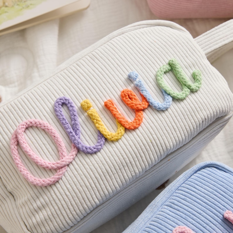 May include: A white corduroy zippered pouch featuring the name Olivia spelled out in colorful, raised rope-style lettering. The letters are pink, purple, yellow, orange, blue, and green, stitched onto the textured fabric surface.