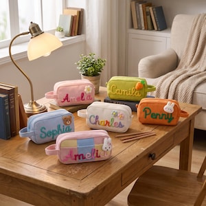 May include: Six rectangular corduroy pencil cases in various colors, each featuring a name embroidered in cursive: Amelia, Camila, Penny, Sophia, Charles, and Amelia. The cases are decorated with small plush appliques like flowers, a bear, a carrot, and rabbits.