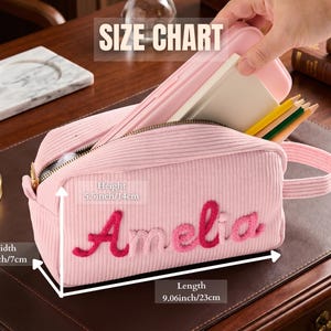 May include: A light pink corduroy pencil case with the name Amelia embroidered in dark pink script. The case features a zipper closure, a side handle, and dimensions of 9.06 inches in length, 2.7 inches in width, and 5.5 inches in height.