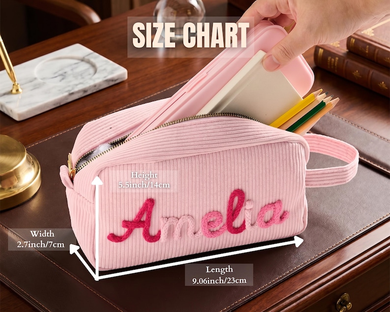 May include: A light pink corduroy pencil case with the name Amelia embroidered in dark pink script. The case features a zipper closure, a side handle, and dimensions of 9.06 inches in length, 2.7 inches in width, and 5.5 inches in height.