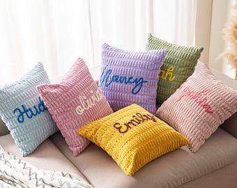 May include: A collection of corduroy throw pillows in various pastel colors, each embroidered with a different name in a complementary color. The pillows are arranged on a neutral-toned sofa, creating a cozy and personalized home decor.
