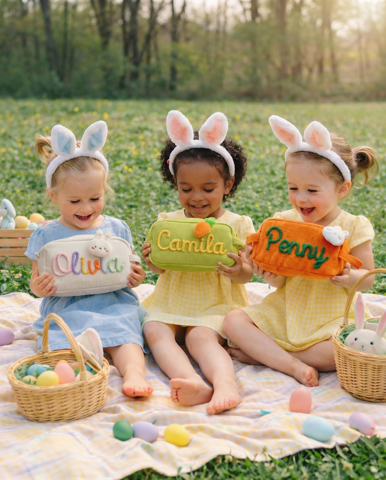 May include: Three rectangular corduroy pouches in white, green, and orange, each featuring embroidered names: Olivia, Camila, and Penny. The pouches are accented with small plush carrot or bunny motifs and are held by children wearing white bunny ear headbands.