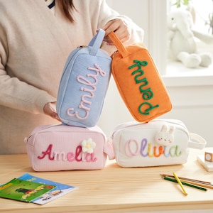 May include: Four rectangular corduroy pouches with handles. One light blue pouch features the name Emily in pink. One orange pouch features the name Penny in green. One pink pouch features the name Amelia and a flower. One white pouch features the name Olivia and a rabbit.