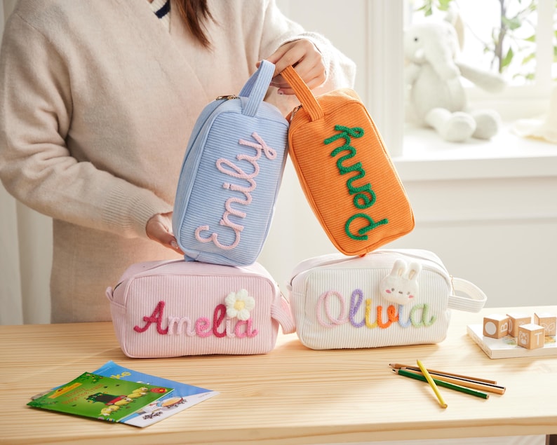 May include: Four rectangular corduroy pouches with handles. One light blue pouch features the name Emily in pink. One orange pouch features the name Penny in green. One pink pouch features the name Amelia and a flower. One white pouch features the name Olivia and a rabbit.