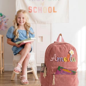 以下が含まれることがあります： フロントポケットにカラフルな糸でAmeliaという名前が刺繍された、ダスティローズ色のコーデュロイ製バックパック。名前の上には小さな白と黄色の花のアップリケがあり、Rusty's Traというタイトルの本がポケットに差し込まれています。
