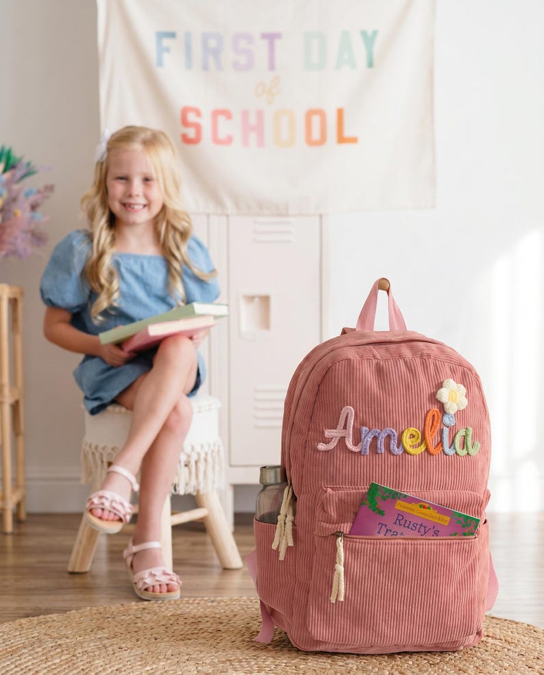 以下が含まれることがあります： フロントポケットにカラフルな糸でAmeliaという名前が刺繍された、ダスティローズ色のコーデュロイ製バックパック。名前の上には小さな白と黄色の花のアップリケがあり、Rusty's Traというタイトルの本がポケットに差し込まれています。