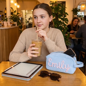 May include: A light blue corduroy zippered pouch featuring the name Emily embroidered in pink script lettering on the front. The rectangular pouch includes a side handle and is positioned on a wooden table next to a tablet and sunglasses.