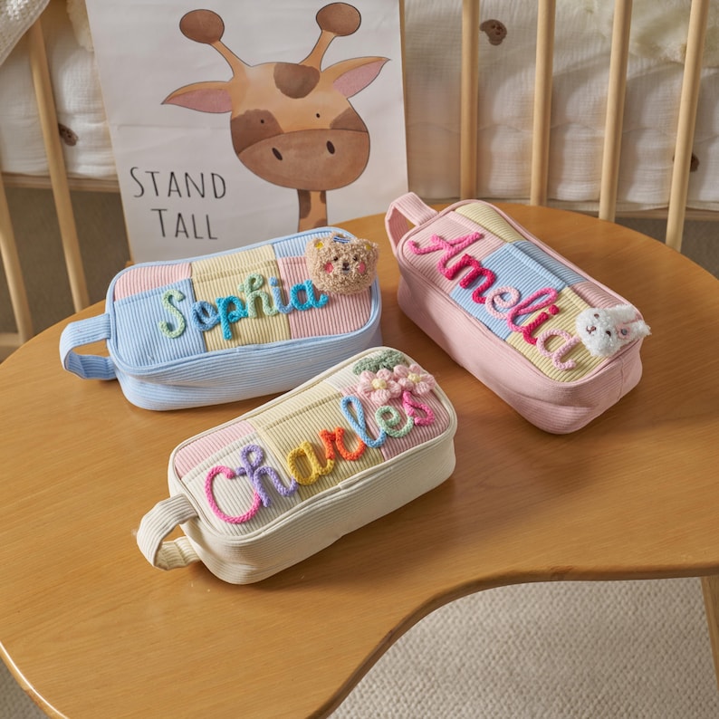 May include: Three rectangular corduroy pencil cases in light blue, pink, and cream. Each case features a colorful embroidered name: Sophia with a bear, Amelia with a rabbit, and Charles with a flower. Each has a side handle.