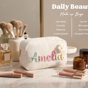 May include: A white corduroy makeup bag featuring the name Amelia embroidered in pastel yarn letters and a small white flower appliqu&eacute;. The bag has a side handle and a zipper closure, displayed with various cosmetic bottles and brushes.