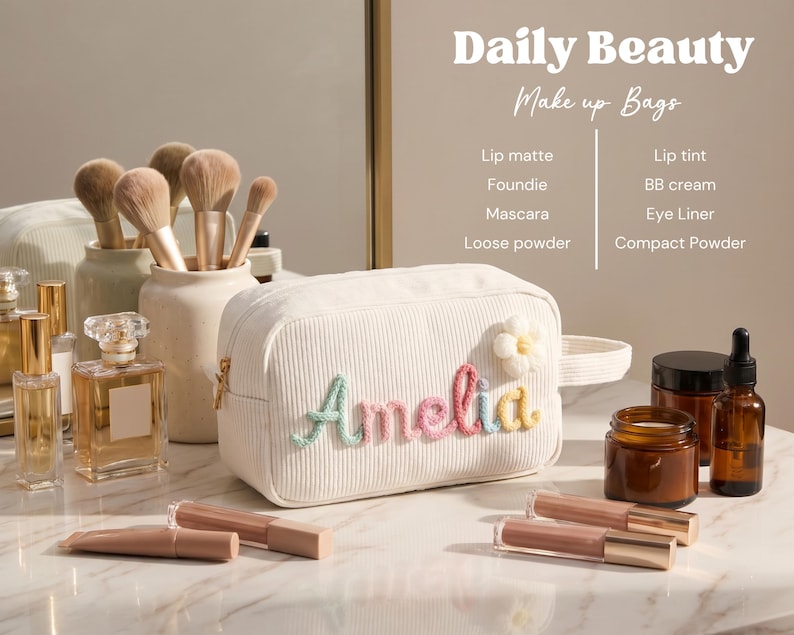 May include: A white corduroy makeup bag featuring the name Amelia embroidered in pastel yarn letters and a small white flower appliqu&eacute;. The bag has a side handle and a zipper closure, displayed with various cosmetic bottles and brushes.
