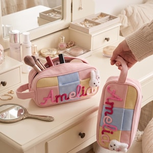 May include: Two rectangular corduroy cosmetic bags in pastel pink, yellow, and blue patchwork patterns. Each bag features the name Amelia embroidered in pink script and a small white plush bunny charm attached to the side zipper pull.