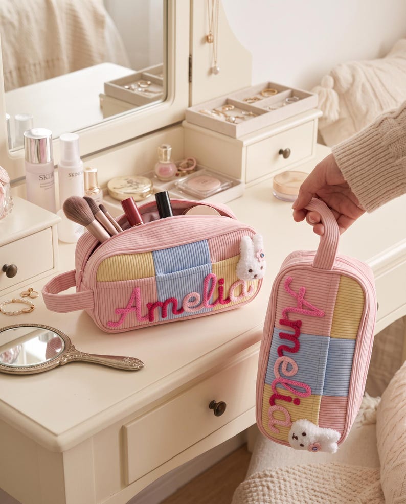 May include: Two rectangular corduroy cosmetic bags in pastel pink, yellow, and blue patchwork patterns. Each bag features the name Amelia embroidered in pink script and a small white plush bunny charm attached to the side zipper pull.