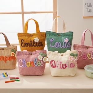 Puede incluir: Una colección de bolsos de mano de pana en varios colores, cada uno bordado con un nombre en una fuente lúdica. Los bolsos tienen asas a juego y adornos florales. También se ve un juego de crayones y un recipiente.