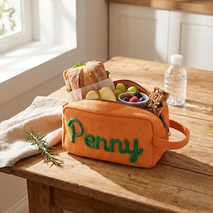 May include: An orange corduroy lunch bag with a handle and a zipper closure, featuring the name Penny embroidered in green cursive script on the front. The bag is filled with a sandwich, apple slices, a small container of berries, and a granola bar.