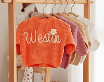 May include: Five colorful knitted sweaters hanging on wooden hangers. The orange sweater has the word "Westin" embroidered on the front with a small white flower.