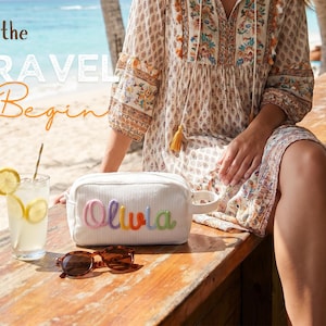 May include: A white corduroy toiletry bag with a side handle and the name Olivia embroidered in colorful yarn letters. The bag is placed on a wooden surface next to a glass of lemonade with lemon slices and a pair of brown sunglasses.