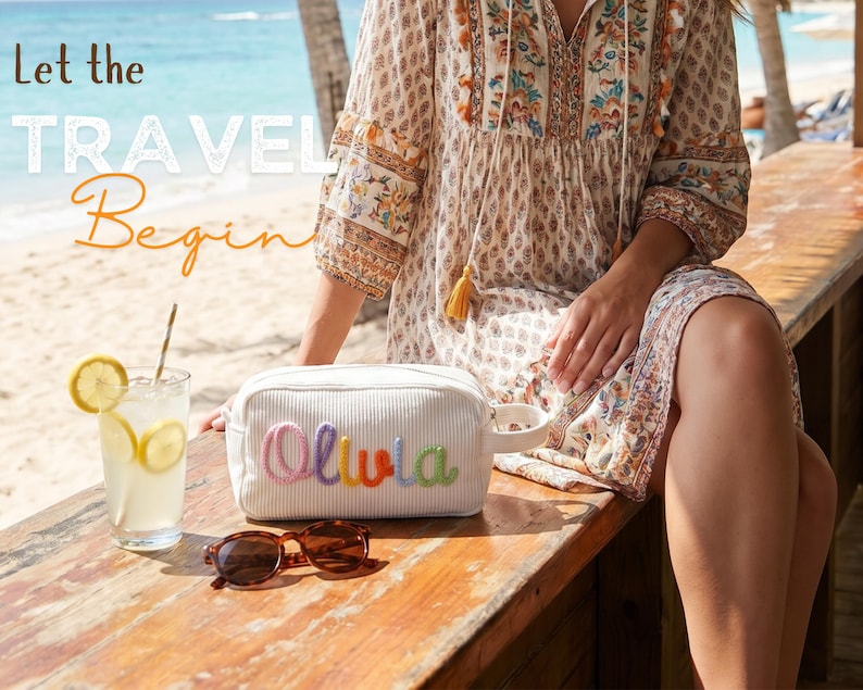 May include: A white corduroy toiletry bag with a side handle and the name Olivia embroidered in colorful yarn letters. The bag is placed on a wooden surface next to a glass of lemonade with lemon slices and a pair of brown sunglasses.