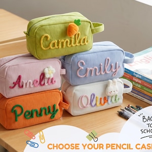 May include: Five rectangular corduroy pencil cases in green, pink, orange, light blue, and white. Each case features a personalized name in raised yarn lettering and a small plush icon, including a carrot, flower, and rabbit.