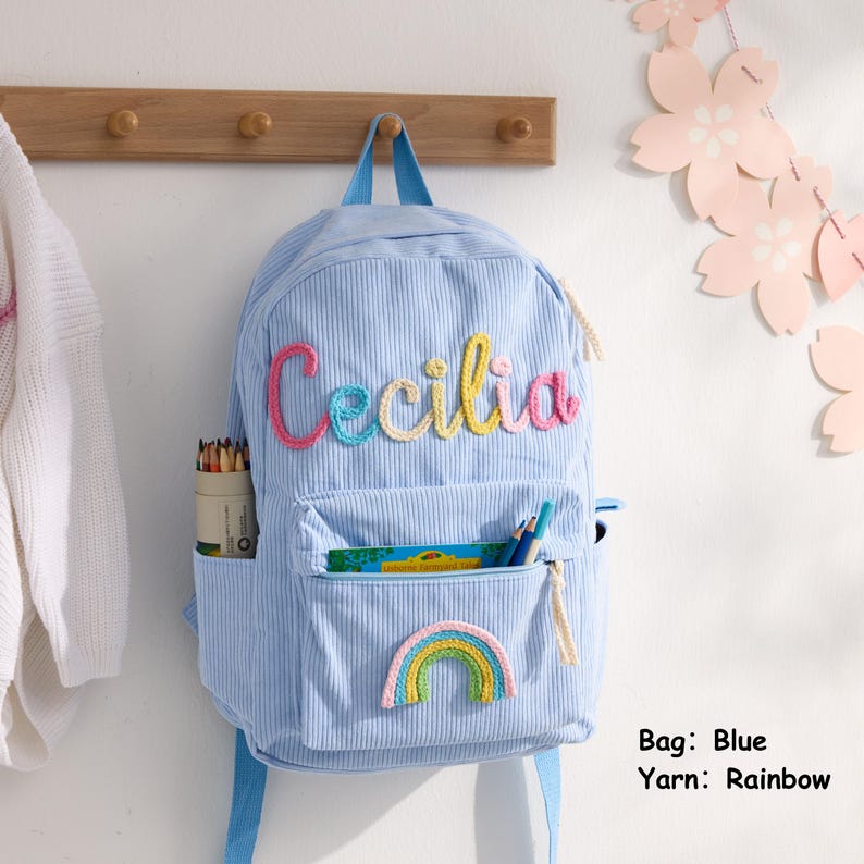 以下が含まれることがあります： 上部にマルチカラーの糸でCeciliaという名前が刺繍された、水色のコーデュロイ製バックパック。フロントポケットには小さな虹のワッペンが縫い付けられています。サイドポケットには色鉛筆が入っています。