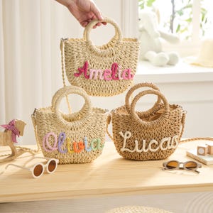 May include: Three woven straw handbags with round handles and shoulder straps. Each bag has a different name embroidered on the front: Amelia in pink, Olivia in rainbow colors, and Lucas in white. The bags are on a wooden surface.
