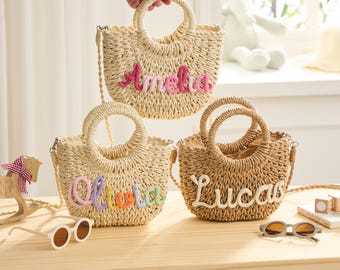 May include: Three woven straw handbags with round handles and shoulder straps. Each bag has a different name embroidered on the front: Amelia in pink, Olivia in rainbow colours, and Lucas in white. The bags are on a wooden surface.