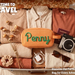 May include: A suitcase packed with a beige sweater, a cream button-down shirt, a rust-colored top, a beige scrunchie, a white charging cable, a passport, a camera, sunglasses, and an orange corduroy pouch with the green embroidered name Penny.