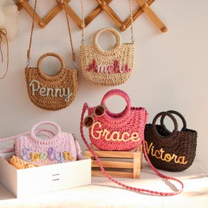May include: A collection of woven straw handbags in various colors, including brown, pink, and black. Each bag features a round handle and a shoulder strap. The bags are personalized with names like "Penny," "Amelia," "Grace," "Evelyn," and "Victoria."