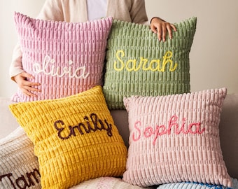 May include: A collection of corduroy throw pillows in various colors, each embroidered with a different name. The pillows are in shades of pink, yellow, and green, with names like "Olivia," "Sarah," and "Emily" stitched in contrasting thread.