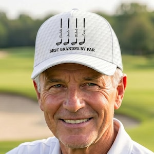 May include: A white golf hat with a diamond pattern and the text "Best Grandpa by Par" is worn by a smiling man. The hat features golf club graphics and names. He wears a white polo shirt and is on a golf course.