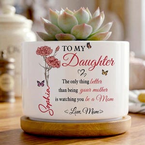 Peut inclure: Un pot de fleurs en céramique blanche avec une plante succulente, portant l'inscription "To My Daughter" et un message affectueux. Le pot repose sur une base en bois et est orné de motifs floraux et de papillons.