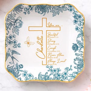 May include: Square, white ceramic trinket dish with a gold rim and teal floral design. The dish features a gold cross and the name "Charlotte" in gold script. Text includes "Beautiful", "Strong", "Enough", "Never Alone", and "Always Loved".