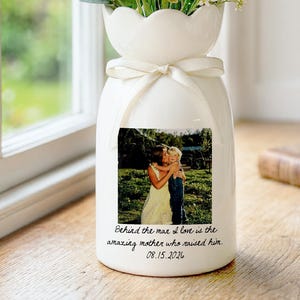 May include: White ceramic vase with a scalloped top, tied with a cream ribbon, filled with yellow and white flowers. A printed photo and text are on the front of the vase. The text reads: "Behind the man I love is the amazing mother who raised him. 08.15.2026."