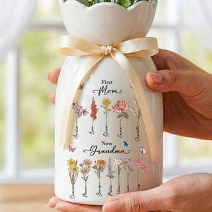 May include: A white ceramic vase with a scalloped top, tied with a beige ribbon. The vase is decorated with floral illustrations and the words "First Mom, Now Grandma" along with names. The vase holds a bouquet of white and yellow flowers.
