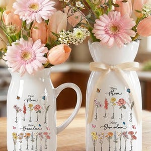 May include: Two white ceramic vases with floral arrangements. The vases are decorated with the words "First Mom, Now Grandma" and floral illustrations. One vase has a handle. The flowers are pink, peach, and white.