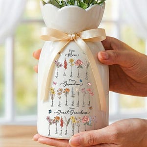 May include: A white ceramic vase with a scalloped top, filled with white and yellow flowers. The vase is decorated with a beige ribbon and features floral illustrations and the text "First Mom, Then Grandma, Now Great Grandma."