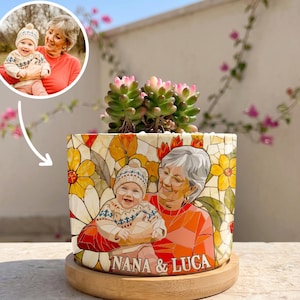 May include: A cylindrical ceramic planter featuring a colorful mosaic-style illustration of an adult and a baby, with the text NANA & LUCA printed at the bottom. The planter holds a green succulent and sits on a circular wooden base.