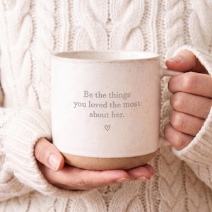 May include: A speckled off-white ceramic mug with a smooth finish and a natural clay-colored base. The front features the gray text "Be the things you loved the most about her." followed by a small hand-drawn heart symbol centered below the text.