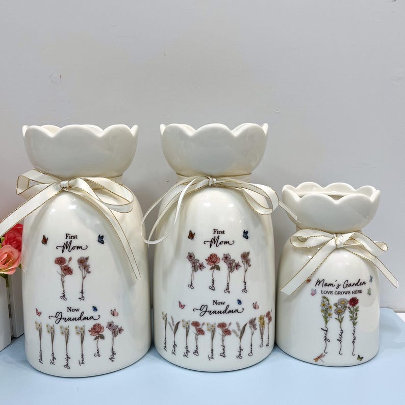 May include: Three white ceramic vases with scalloped tops and cream-colored ribbon bows. The vases are decorated with floral designs, butterflies, and text that reads "First Mom, Now Grandma" and "Mom's Garden LOVE GROWS HERE."