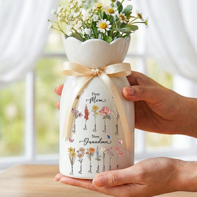 May include: A white ceramic vase with a cream-colored ribbon tied around the neck, holding a bouquet of white and yellow flowers. The vase is decorated with floral illustrations and the words "First Mom, Now Grandma."