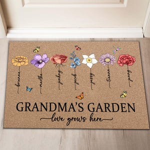 May include: A brown doormat with the text "Grandma's Garden - love grows here" and a row of colorful flowers with names written underneath each flower. The names are Brianna, Martha, Johnny, Jacob, Jassy, Tianna, and Jenny.