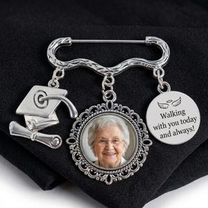 May include: Silver-toned graduation brooch with a photo pendant. The brooch features a mortarboard and scroll charm, a circular pendant with a photo, and a charm with the text "Walking with you today and always!"