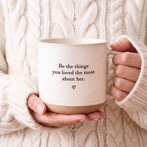 May include: A speckled off-white ceramic mug with a smooth, unglazed tan base. The front features black printed text that reads, Be the things you loved the most about her, followed by a small hand-drawn heart symbol centered below the text.
