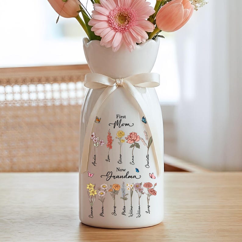 May include: A white ceramic vase with a cream-colored ribbon tied around the neck, holding pink and peach flowers. The vase is decorated with floral illustrations and the words "First Mom Now Grandma" with names.