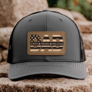 May include: A grey trucker cap with a black mesh back and a curved peak. The cap has a brown leather patch with the word "DAD" in a wood-grain design, with the American flag stars and stripes. The names "ANNA HENRY WILLIAM" are below.