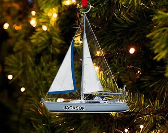 May include: A sailboat ornament with white sails and a blue trim, suspended by a red ribbon. The boat is light gray with the name "JACKSON" printed on the side. The background is a blurred Christmas tree with lights.