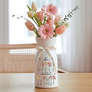 May include: A white ceramic vase with a ribbon, filled with pink gerbera daisies, tulips, and other flowers. The vase has the words "First Mom New Grandma" and names printed on it. The vase is on a wooden table.
