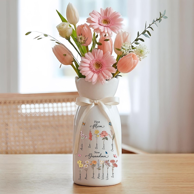 May include: A white ceramic vase with a ribbon, filled with pink gerbera daisies, tulips, and other flowers. The vase has the words "First Mom New Grandma" and names printed on it. The vase is on a wooden table.