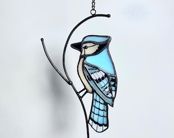 May include: Stained glass blue jay bird suncatcher hanging from a black metal branch and chain. The bird features light blue, white, and black details. The suncatcher is designed to hang in a window or on a wall as a decorative piece.